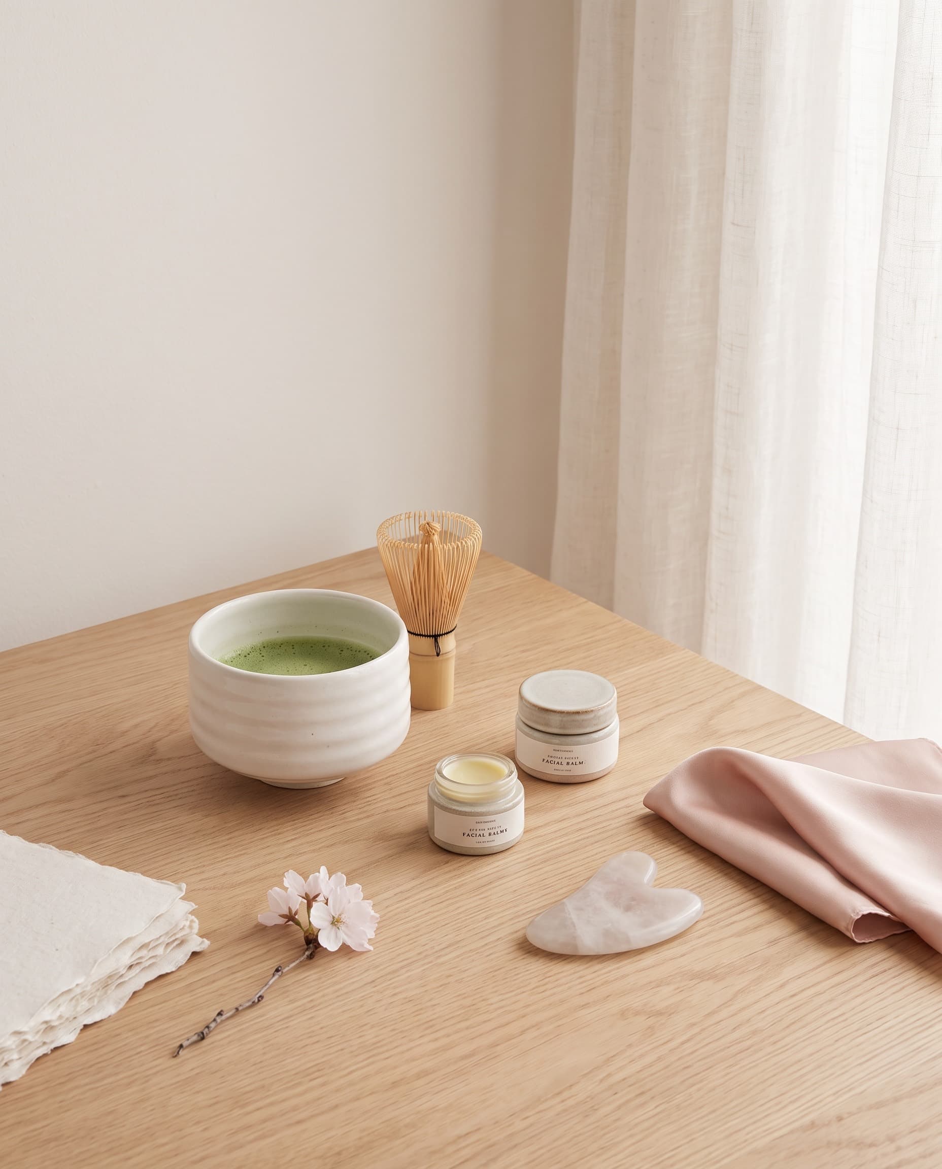 Matcha bowl with a bamboo whisk beside facial balms and a sakura blossom